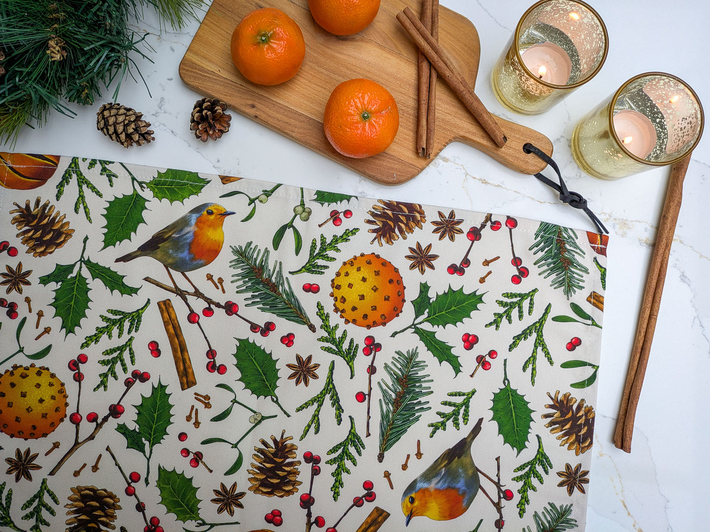 Close up of colourful Christmas tea towel with lit candles, clementines, pine cones, cinnamon sticks and marble worktop