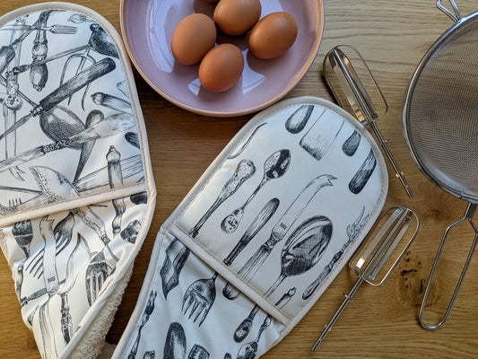Beautifully detailed cutlery illustrated organic cotton oven gloves shown with baking utensils and fresh eggs ready for baking