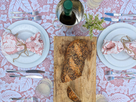 Dining table setting with fresh bread on a wooden board, floral pastel fresh tablecloth, and cutlery.