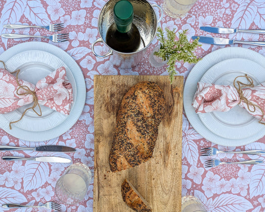 Dining table setting with fresh bread on a wooden board, floral pastel fresh tablecloth, and cutlery.