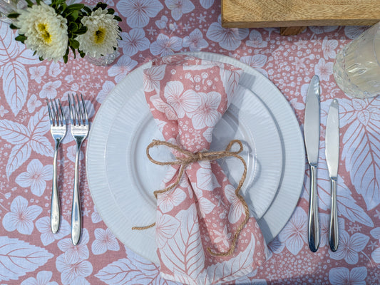 Inviting dinner setting with white plates, silverware, and a soft pink hydrangea floral tablecloth and napkin