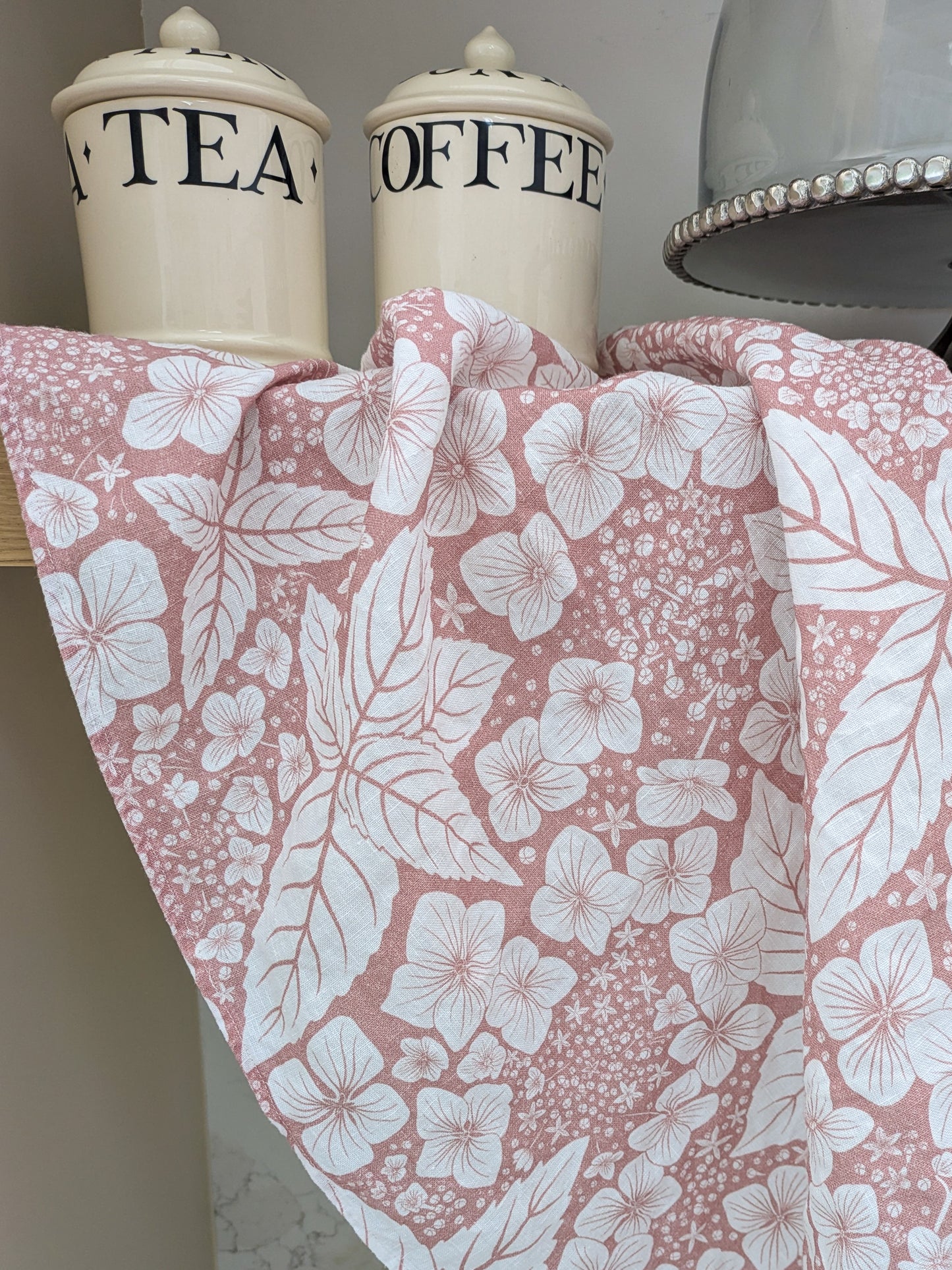 Pink floral tea towel draped on a shelf with 'Tea' and 'Coffee' jars and a silver cake stand