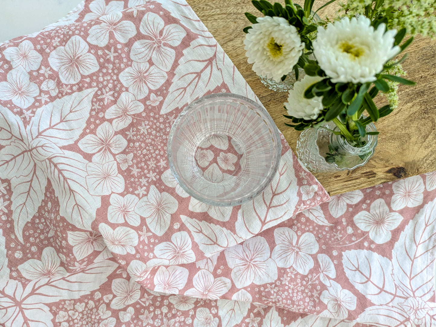 Floral-patterned pink tea towel with a glass and white flowers in bud vases on a wooden surface