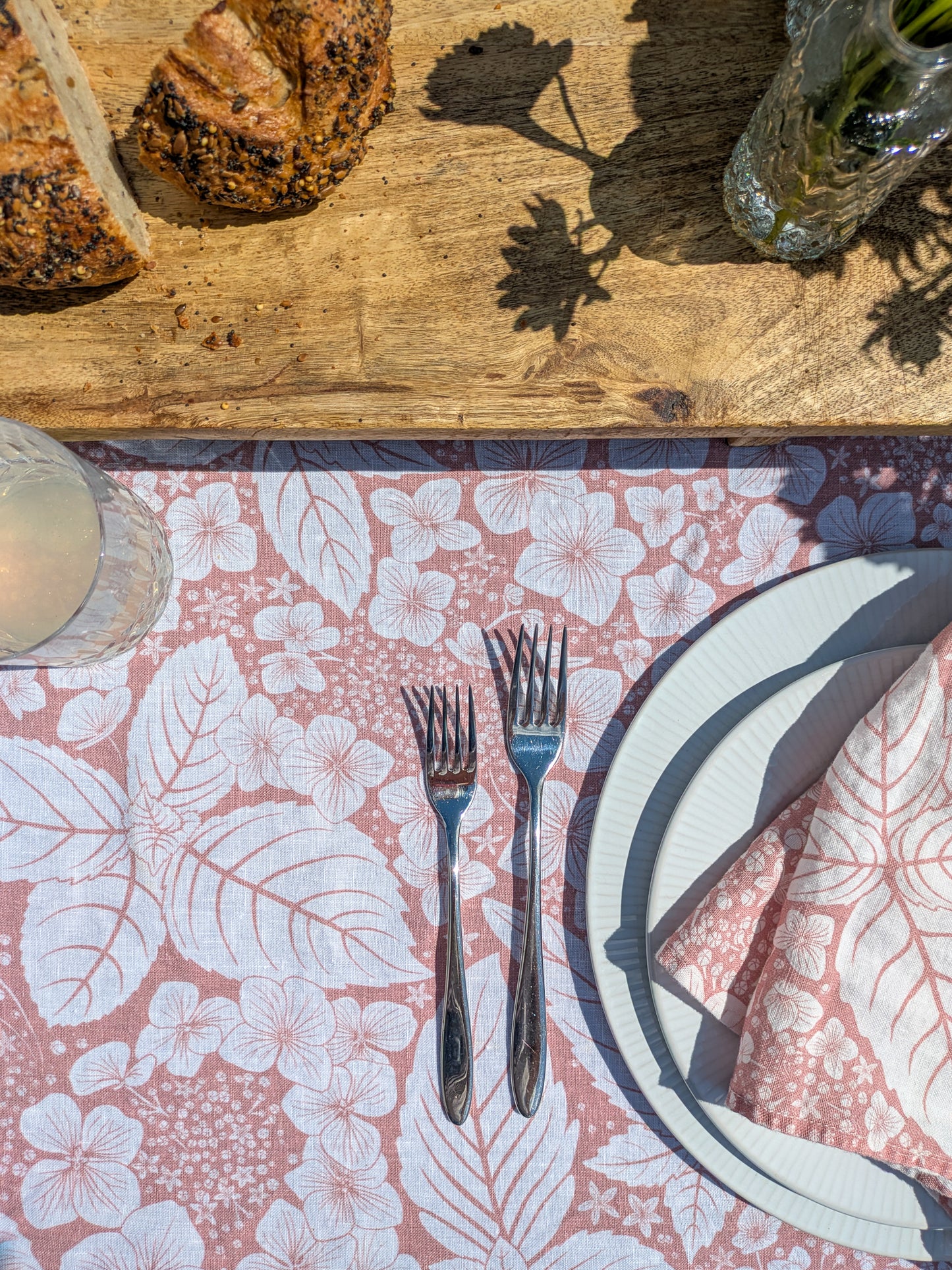 Table setting with pink hydrangea tablecloth, cutlery, bread on a wooden surface and cut glass bud vases filled