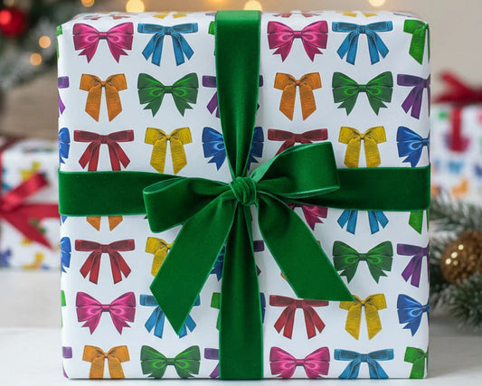 A square gift box wrapped in white paper printed with colorful bow motifs, tied with a large green ribbon and bow, set against a softly blurred holiday background with warm lights and a Christmas tree.