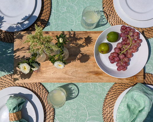 Table setting with the delicate green tomato tablecloth including plates, full glasses, fresh cut flowers, fruit and warm sunlight shining