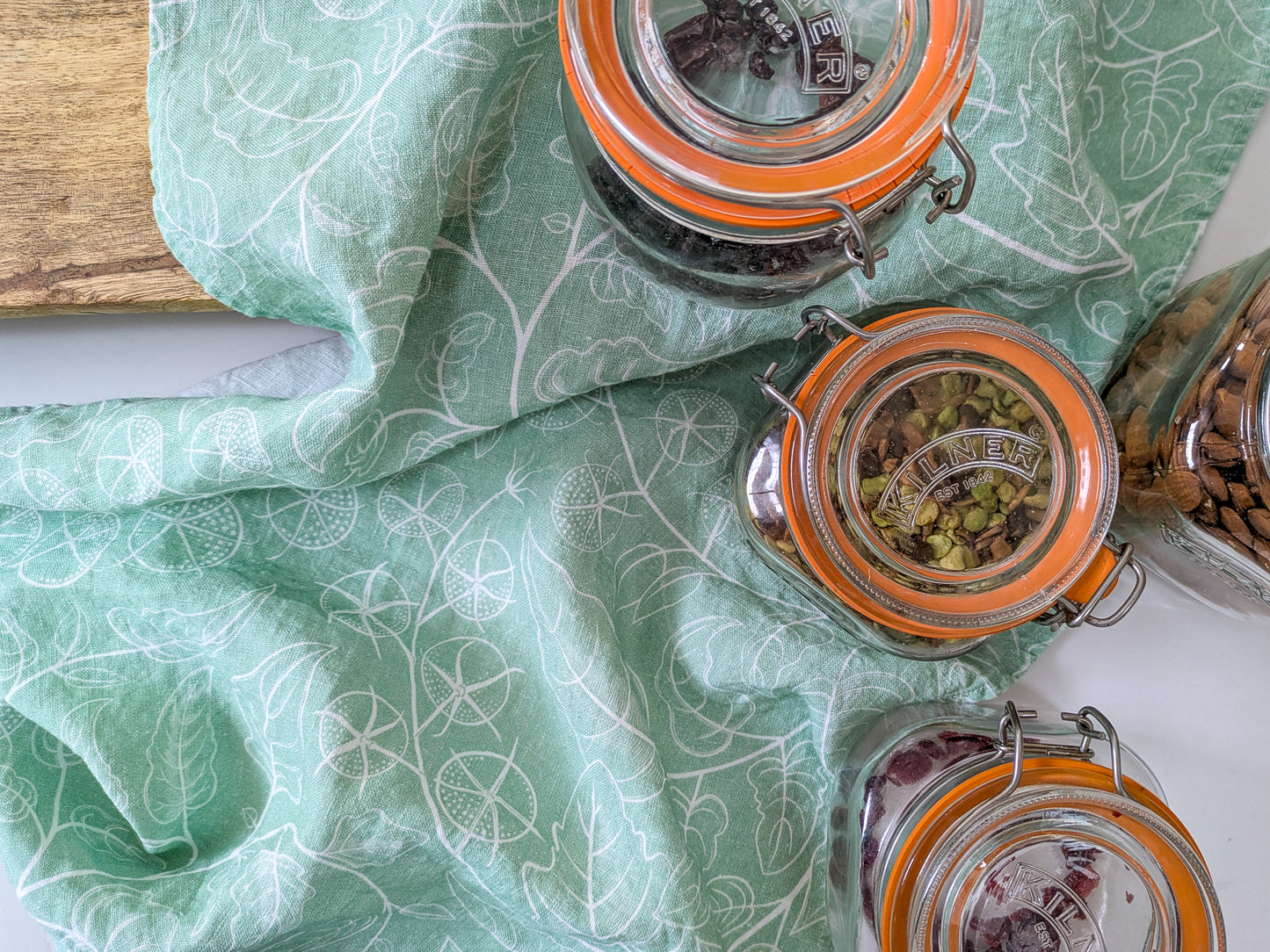 Kilner jars full of nuts and pulses with tomato soft green patterned tea towel on a marble worktop with oak board