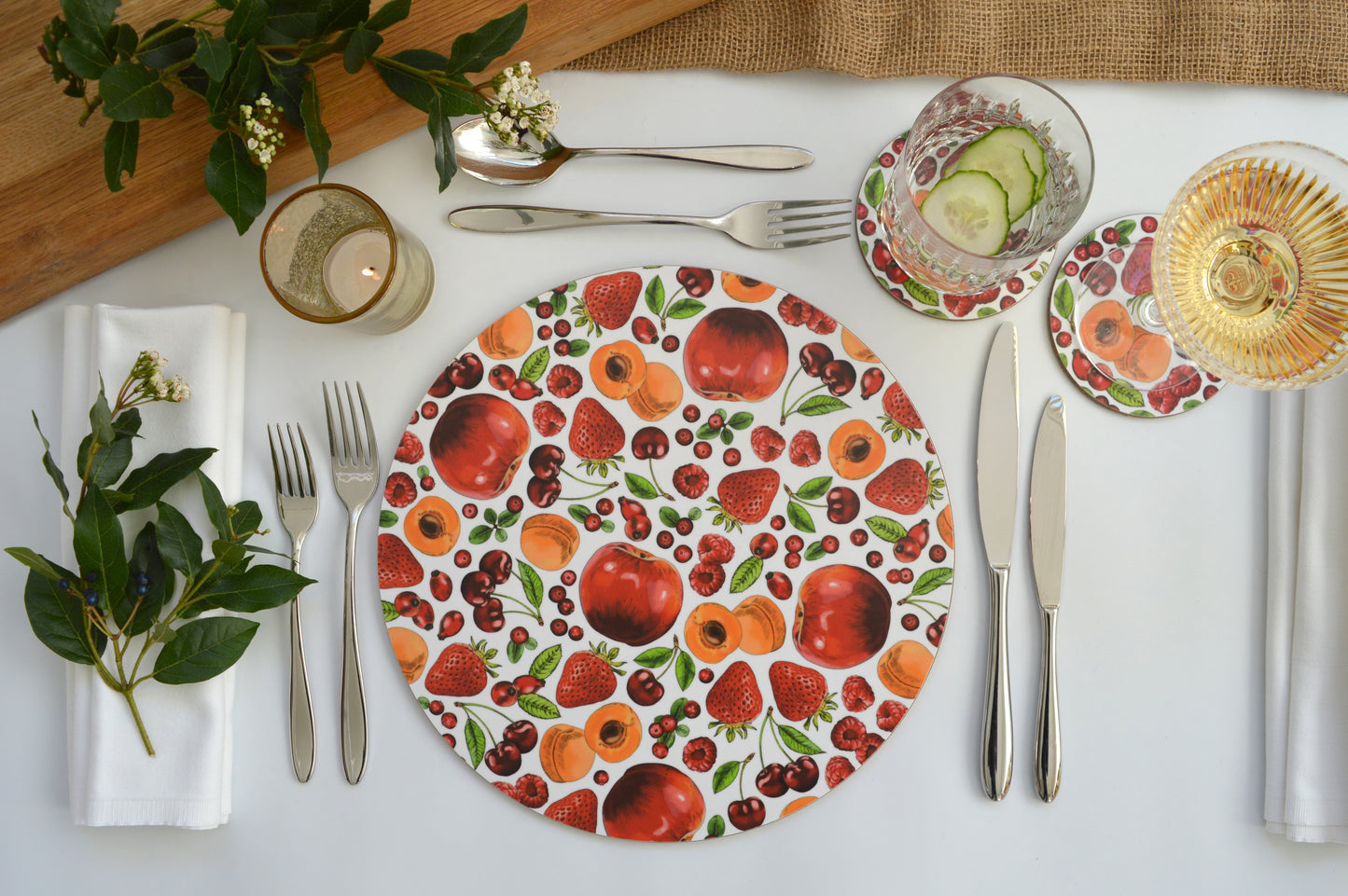 Red and orange fruit illustrated coasters pictured with matching placemat on an inviting laid table with fresh foliage and poured drinks