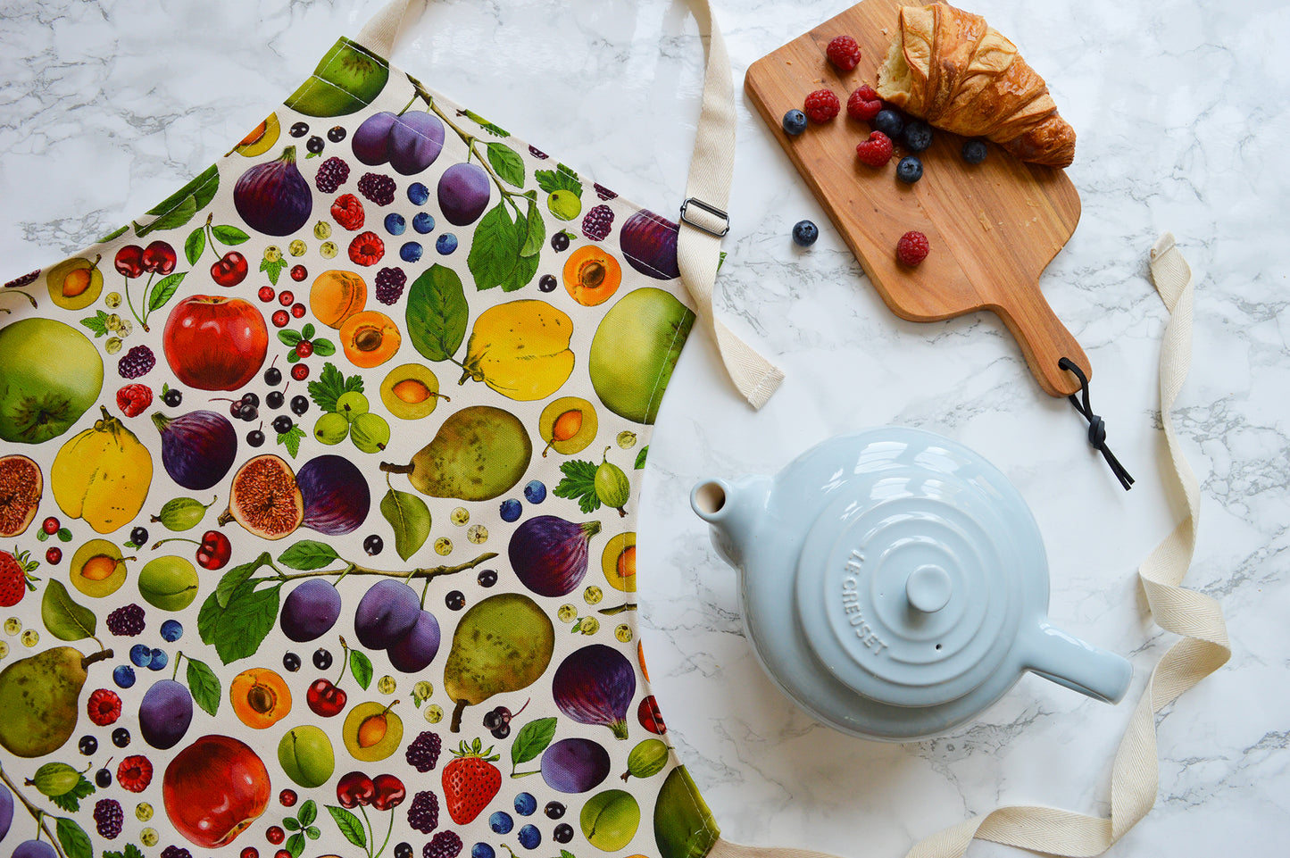 Flat lay of British colourful fruit patterned apron with fresh berries, croissant and warm tea