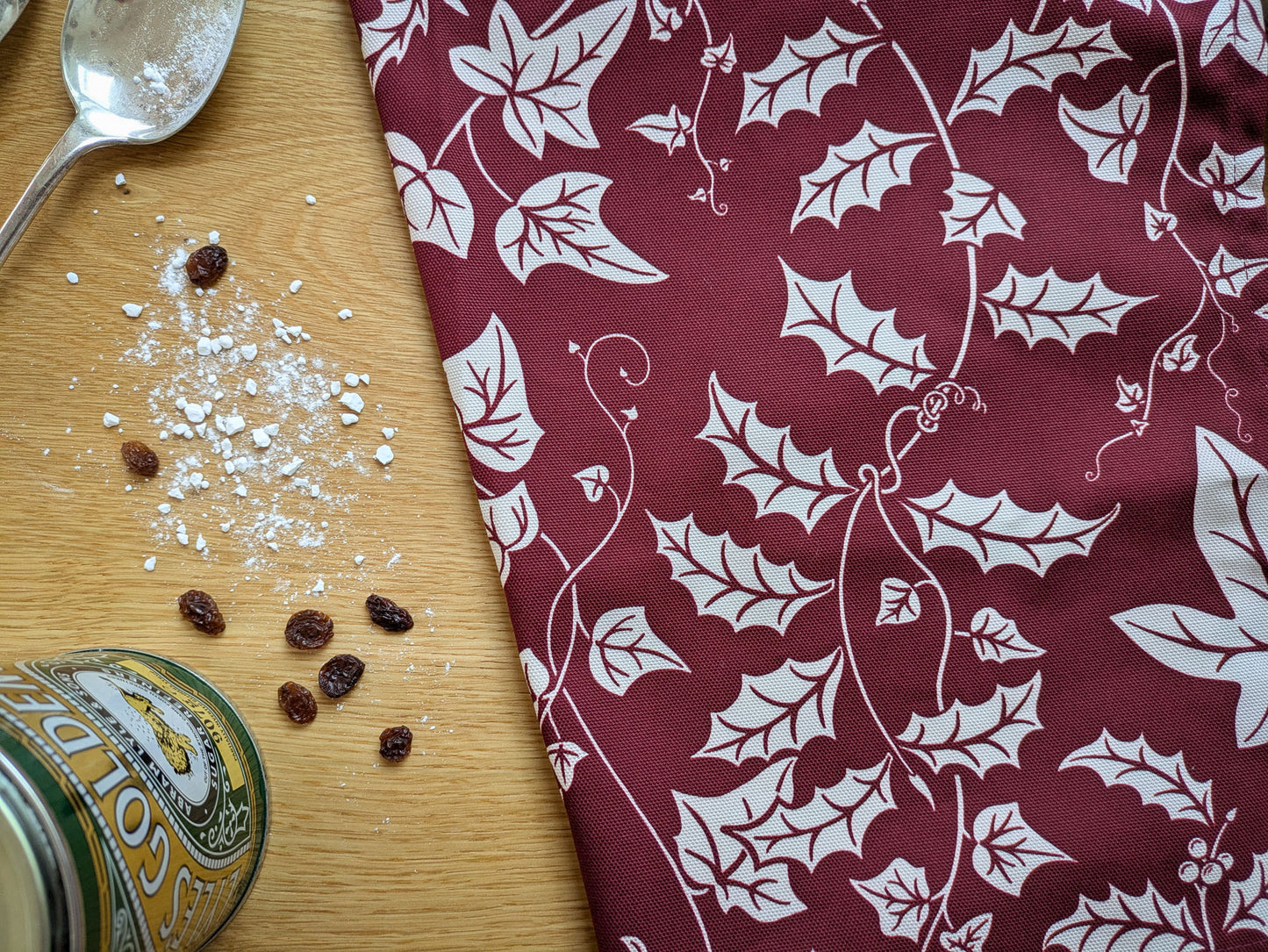 Close up of the burgundy Holly and Ivy illustrated tea towel with baking ingredients on an oak worktop