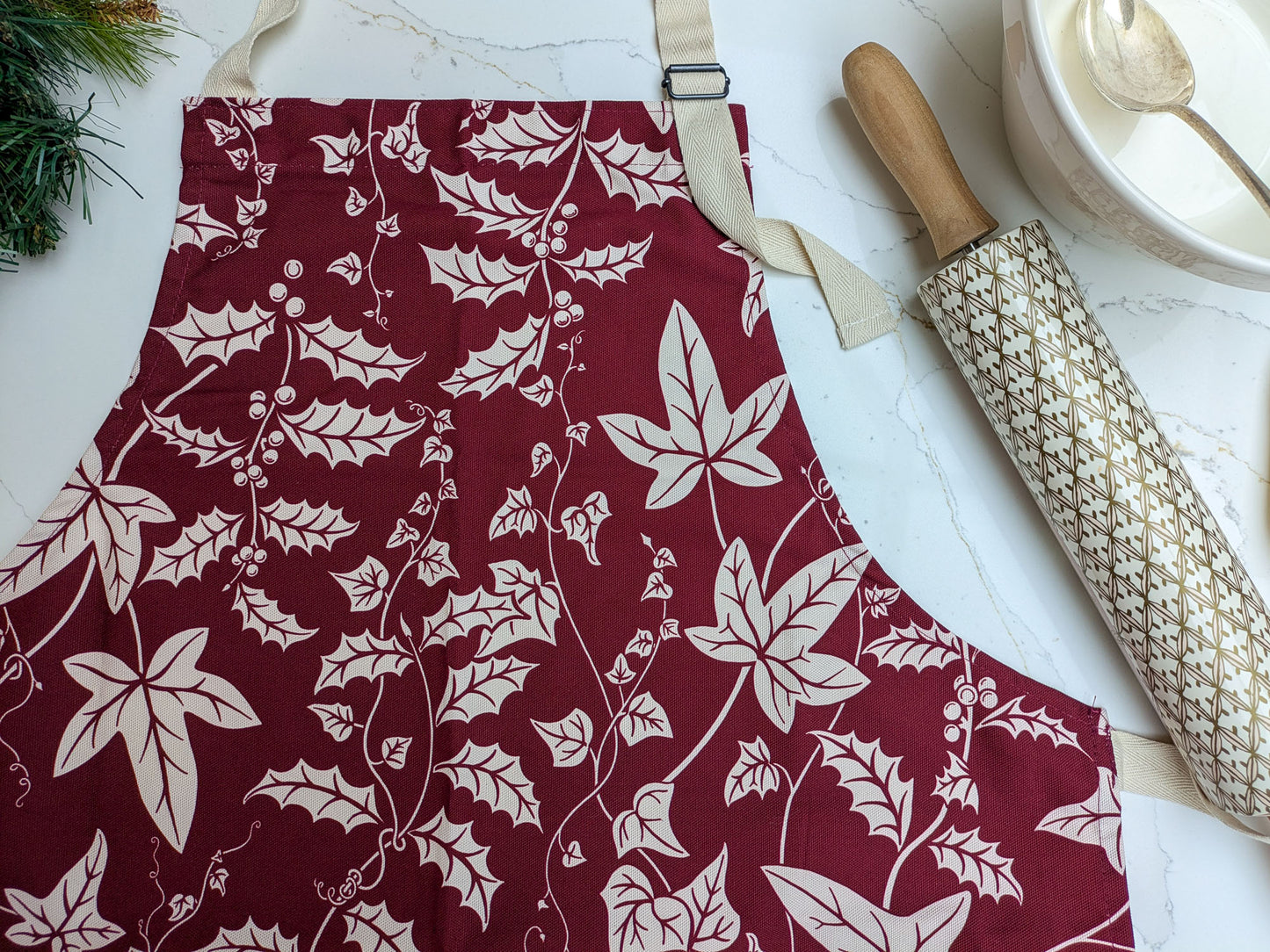 Holly and Ivy Botanical illustrated kitchen apron on a marble surface with patterned rolling pin and baking equipment