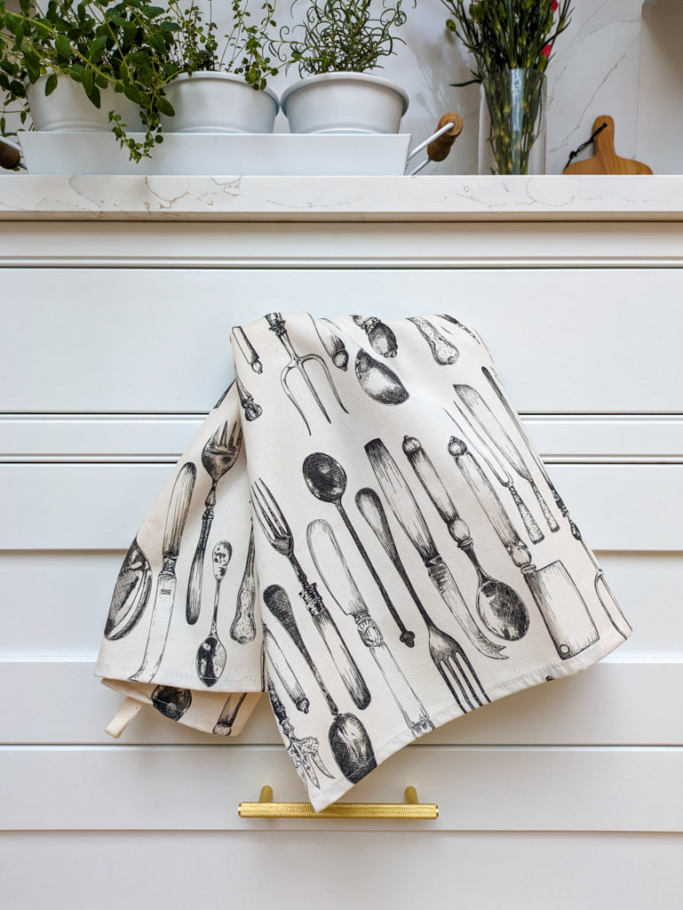 Detailed natural cotton tea towel illustrated with vintage cutlery in a crisp white kitchen with fresh growing herbs