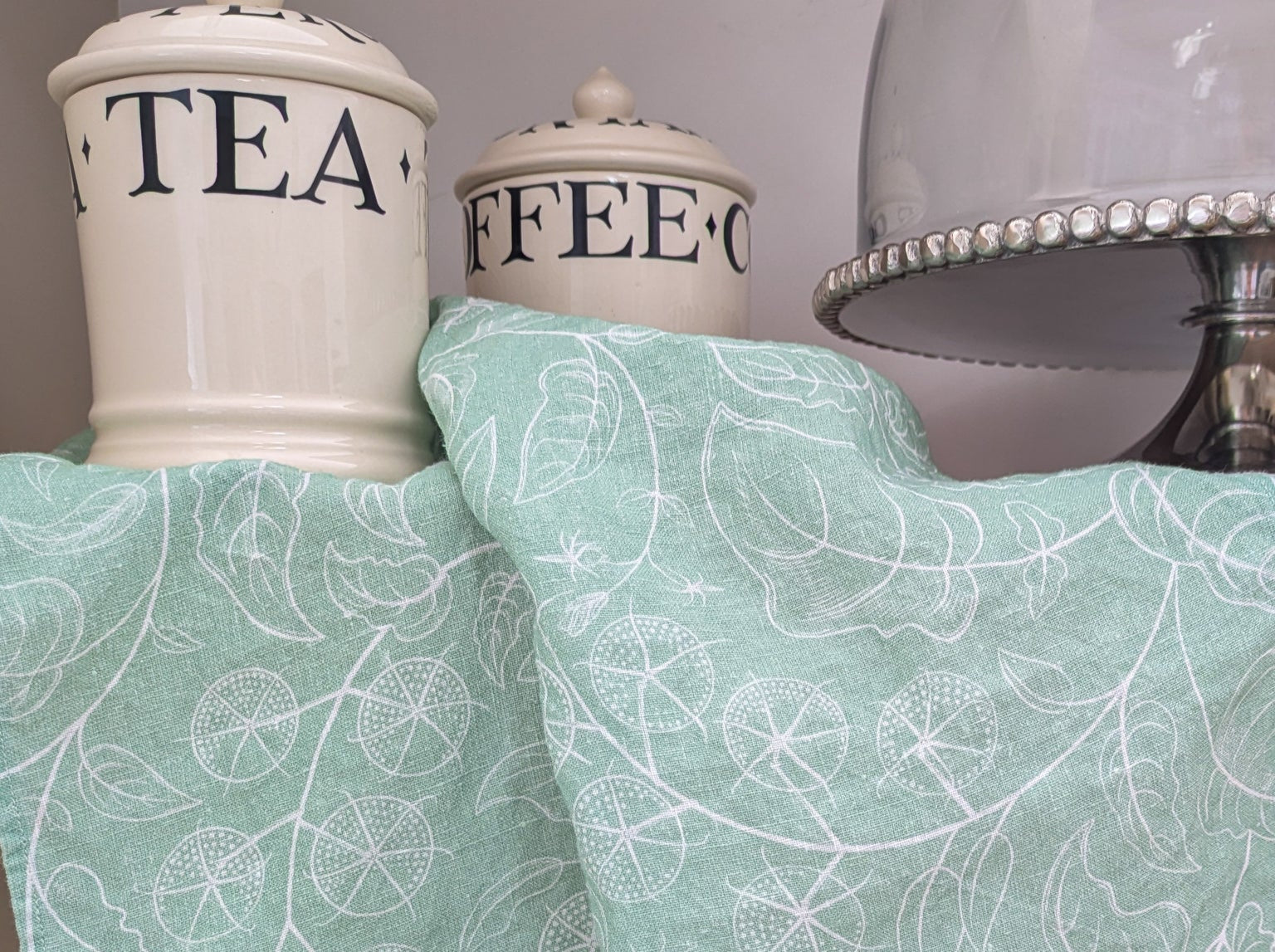 Tea and coffee canisters and a cake stand on shelf with tomato soft green tea towel draped beneath
