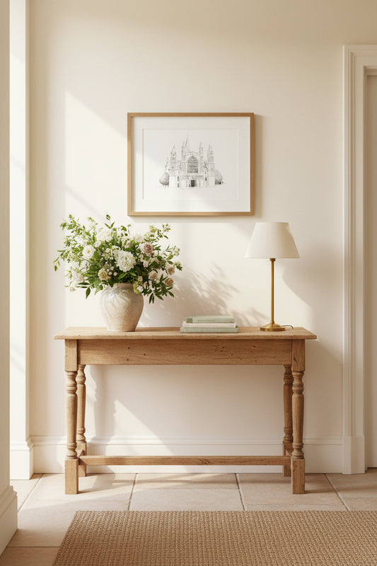 A styled interior scene showing a light wood console table against a cream wall, with a vase of white flowers, a table lamp, and a framed black-and-white architectural print above it of Rochester Cathedral