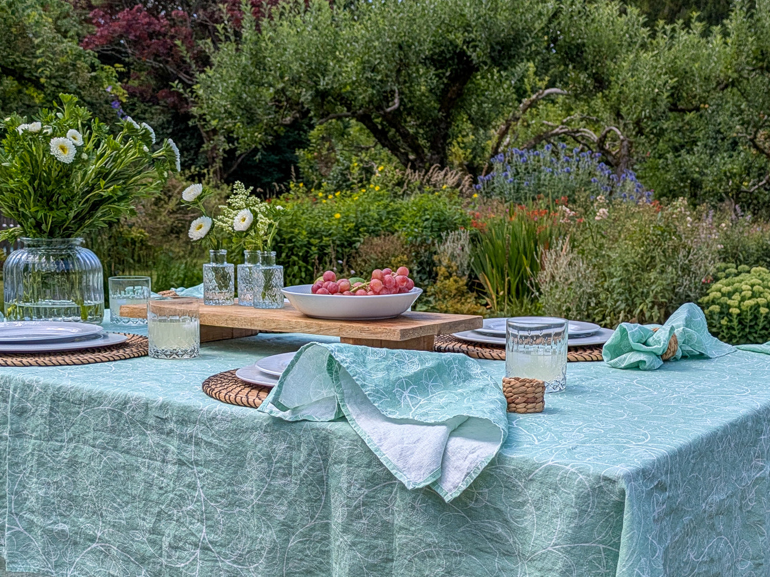 Gorgeous outdoor table setting with a garden view. Soft green tomato table cloth and matching napkins set the scene for an inviting table with fresh fruit and flowers