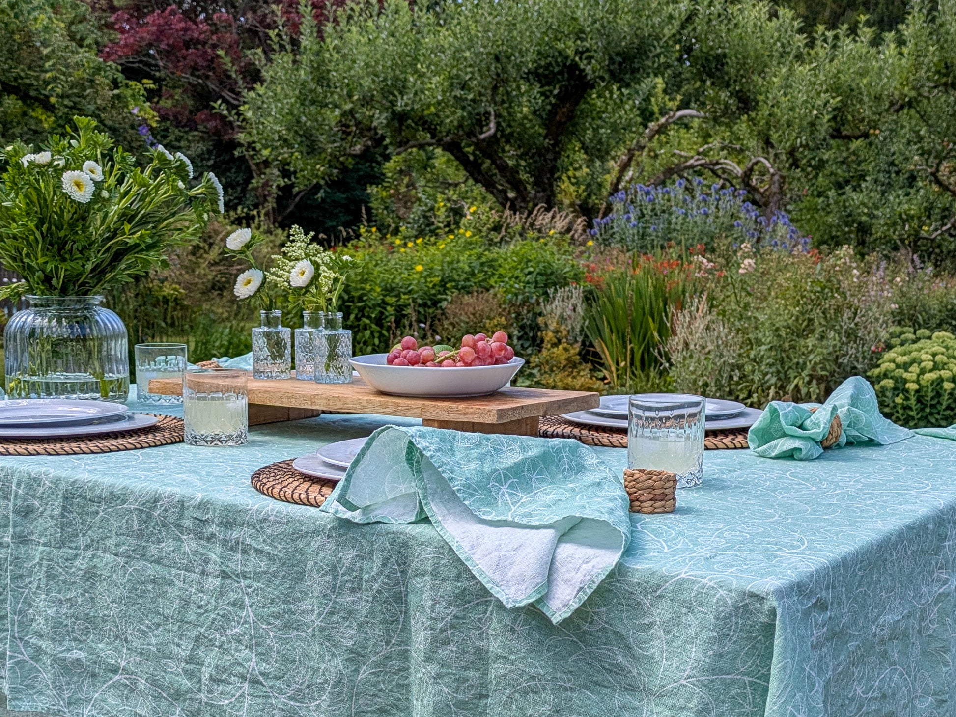 Gorgeous outdoor table setting with a garden view. Soft green tomato table cloth and matching napkins set the scene for an inviting table with fresh fruit and flowers
