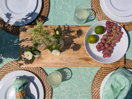 Table setting with the delicate green tomato tablecloth including plates, full glasses, fresh cut flowers, fruit and warm sunlight shining