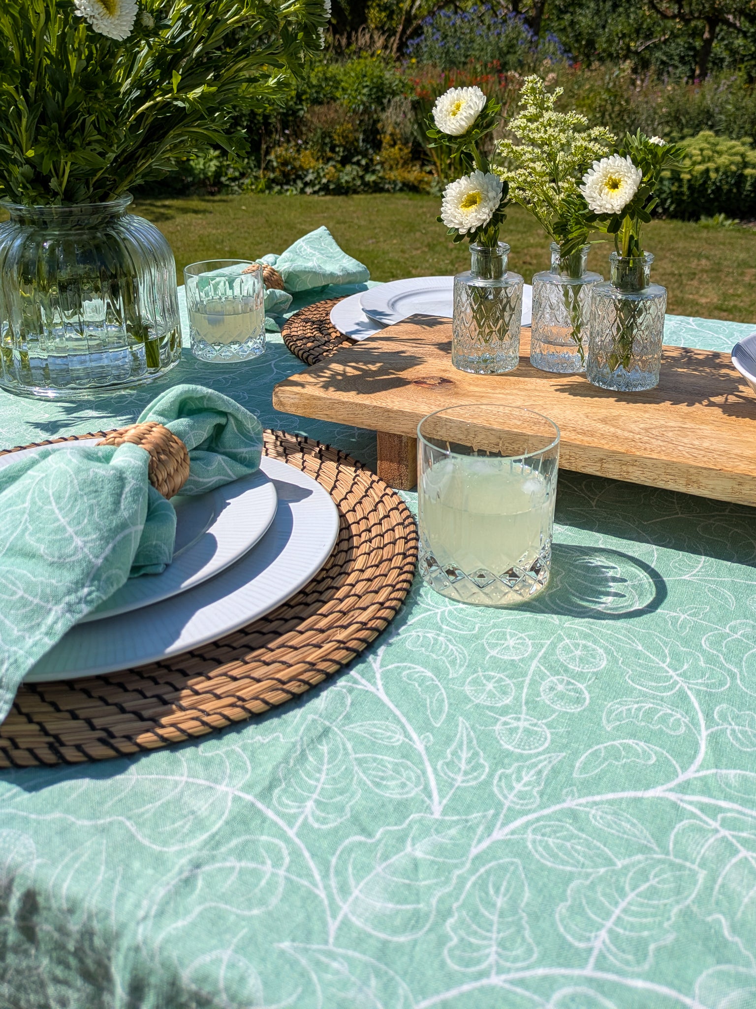 Outdoor table setting with green tomato tablecloth, full glasses, floral arrangement and inviting setting