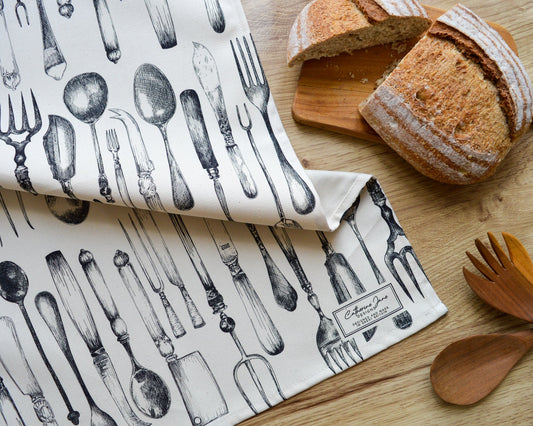 Knife, fork and spoon tea towel with freshly baked bread on a warm wooden worktop